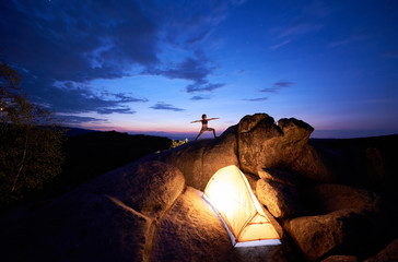 Camping on rock formation. Brightly lit tourist tent and silhouette of slim tourist girl doing yoga exercises on mountain top against dark blue sky at sunset. Sport, tourism concept. Virabhadrasana