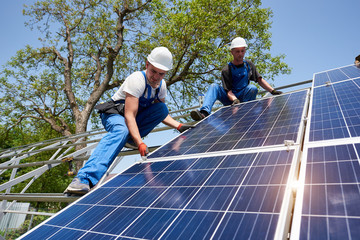 Two young technicians installing heavy solar photo voltaic panel on tall steel platform on green...