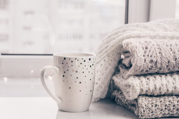 Fototapeta na wymiar Stack of warm cozy knitwears and a cup of coffee on white marble windowsill against white window background. Copy space.
