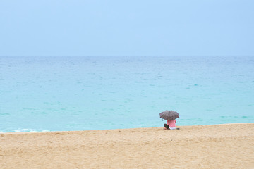 A woman under umbrella on the sandy beach looking at the water under cloudy sky on a rainy day.