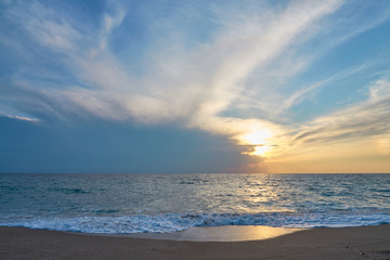 Fototapeta premium Sunset at the tropical beach, sun behind clouds reflects on water and waves with foam hitting sand.