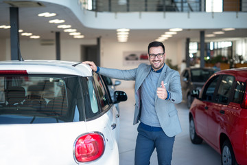 Car buyer with thumbs up standing by the new vehicle in car dealership showroom. Vehicle sale.
