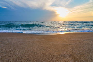 Sunset at the tropical beach, sun behind clouds reflects on water and waves with foam hitting sand.