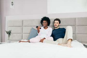 Obraz na płótnie Canvas Happy interracial couple in home wear, relaxing in bed holding remote control and watching television