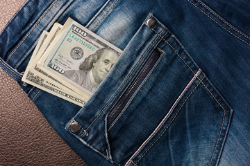 American dollar bills in a jeans pocket are on the leather surface.