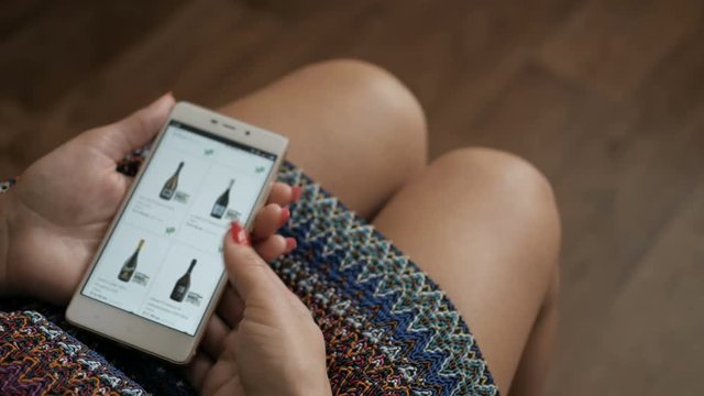 Young woman with manicure orders champagne wine home in an online store. Female selects the alcohol in the grocery online store. Close up. 4K UHD. Screen is blurred.