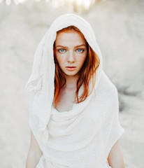 A beautiful young girl with red hair and freckles looks intently at the camera. Woman in the hood and clothes for the desert. Concept - obrazy, fototapety, plakaty