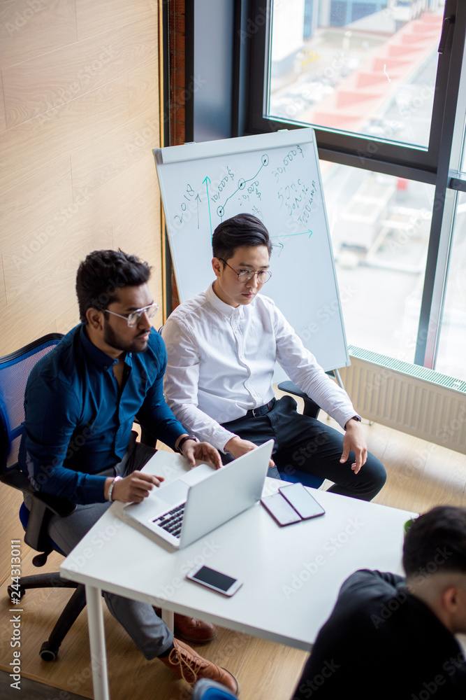 Wall mural image of two young indian and asian colleagues interacting, sitting beside each other in office with