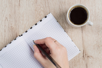 young woman hand hold opened notebook pages with black pen next to cup of coffee on  wooden table. Top view.