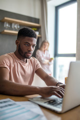African young businessman working at home in his day-off, making presentation at home with laptop all night instead of being together with his beautiful caucasian wife, standing lonely on background