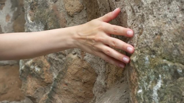 Woman sliding hand against old ancient stone wall in slow motion. Female hand touching hard rough surface of rock with green mold on it