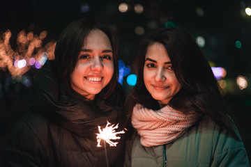 Two positive girlfriends looking at camera on the street with sparkler in hand. Bright bokeh lights of Christmas tree and garlands on background. Winter holidays mood.