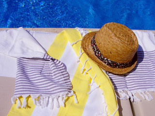 Trendy straw hat with ribbon and cotton towels on a sunbed near the swimming pool. Solar glare on...