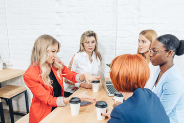 Team work process. Multiracial group of young women collaborating in open space office. Red-haired mentor coach speaking to young people, teaching audience at training seminar.