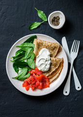 Delicious breakfast or brunch - crepes with smoked salmon, spinach and sour cream on a dark background, top view