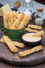 Homemade savory bread sticks with cheese and sesame in a basket, served with sauce on wooden board, vertical