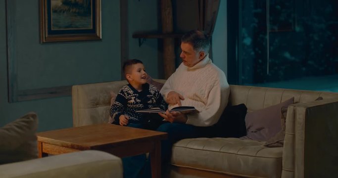 Grandfather reading book with grandson at home near fireplace. 4K UHD 60 FPS SLOW MOTION Blackmagic RAW