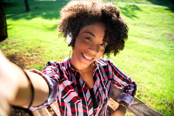 Closeup portrait of smiling young attractive African American woman holding smartphone, taking selfie photo and standing in park with blurred plants in background - Imagem