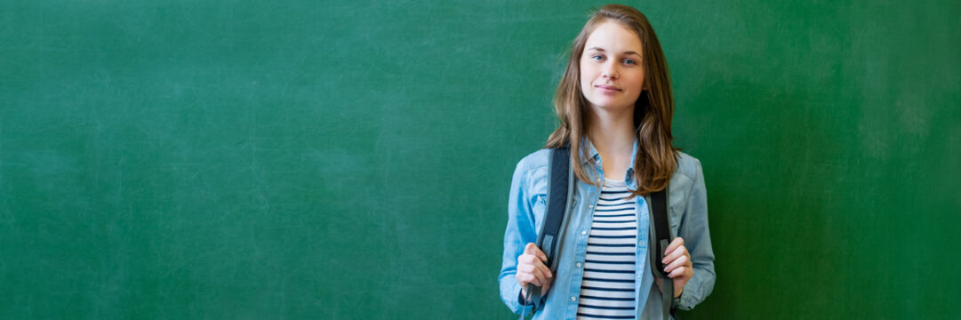 Young Confident Smiling Female High School Student Standing In Front Of Chalkboard In Classroom, Wearing Backpack, Looking At Camera. Education Concept Banner.