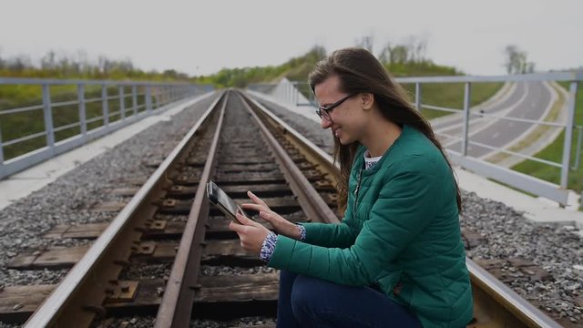 Young smiling girl using tablet and standing at railway. Wearing glasses and dressed in green.
