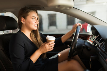 Young gorgeous woman in black dress sitting behind the wheel driving car holding coffee in hand while dreamily looking straight