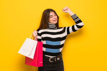 Young woman over yellow wall holding a lot of shopping bags in victory position