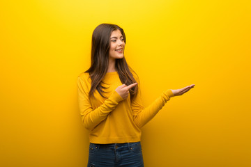 Teenager girl on vibrant yellow background holding copyspace imaginary on the palm to insert an ad