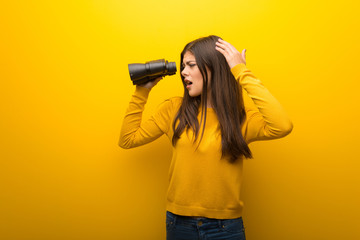 Teenager girl on vibrant yellow background and looking in the distance with binoculars
