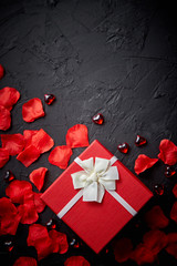 Gift box on black stone table. Sweet romantic holiday background with rose petals, acrylic heart shaped decoration. Valentines day background. Top view with copy space.