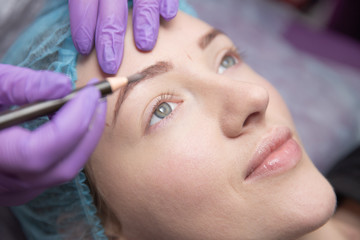 Permanent make-up for eyebrows of beautiful woman with thick brows in beauty salon. Closeup beautician doing tattooing eyebrow.