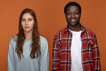Picture of handsome positive young dark skinned male smiling, standing next to emotional white girl with long loose hairstyle who is staring at camera with frightened bug eyed facial expression