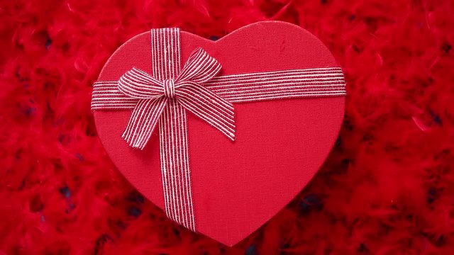 Heart shaped boxed gift, placed on red feathers background. Valentines day or Romantic date concept. Top view shot. With copy space