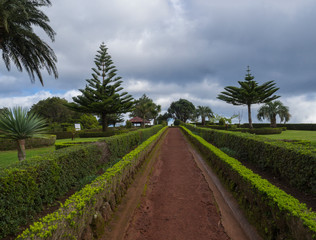 Fototapeta na wymiar Dirt footpath in gardens at viewpoint Ponta do Sossego with green bush, palm trees and altan gazebo, Sao Miguel Island, Azores, Portugal