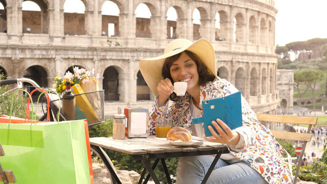 Happy young woman tourist taking selfies with smartphone at the table outside a bar restaurant in front of the Colosseum in Rome with coffee, juice and cornetto. Elegant beautiful dress with large hat