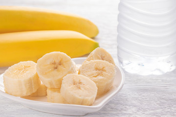 Sliced and whole bananas. On a white plate. On the old wooden table. Misted glass with clean water