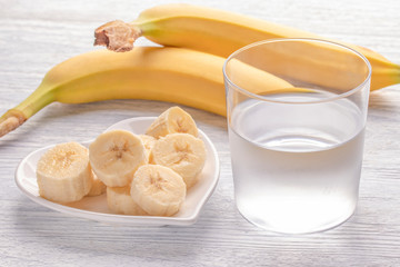Sliced and whole bananas. On a white plate. On the old wooden table. Misted glass with clean water