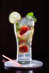 Freshly made refreshing mojito in a tall glass with strawberry, lime, fragrant mint leaves and ice on a dark background next to the color cocktail tubes