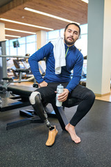 Portrait of young disabled man with amputee leg sitting with bottle of water and resting after sport training in gym