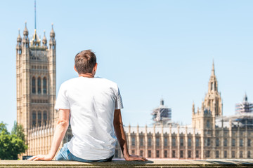 Back of young man with white tshirt sitting on railing in London looking at cityscape skyline of...