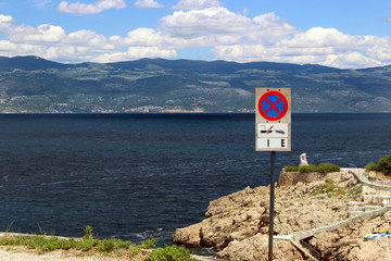 No parking and tow away sign directly by the Adriatic Sea Bay with the opposite shore and blue cloudy sky as background