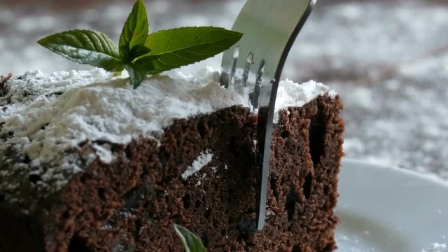 Homemade baked chocolate brownie cake muffled with powdered sugar on a white plate decorated with mint leaves. Fork breaks off piece of brownie pie from the plate