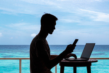 silhouette of a young man working with a computer and a smartphone on a table. Clear blue tropical water as background