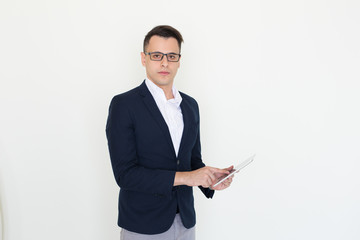 Serious young businessman studying report on tablet. Handsome Caucasian man in glasses and jacket using mobile computer and looking at camera. Business and communication concept