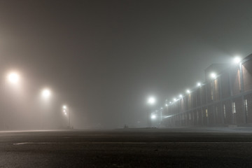 Fototapeta premium Empty urban car parking and streetlights at foggy night. Old Industrial brick building and lanterns on lonely street