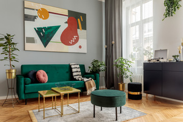 Luxury and modern home interior with design green sofa, navy commode, tables, pouf and accessroies. A lot of plants in the room. Abstract painting.  Stylish decor of living room with brown parquet.  - obrazy, fototapety, plakaty