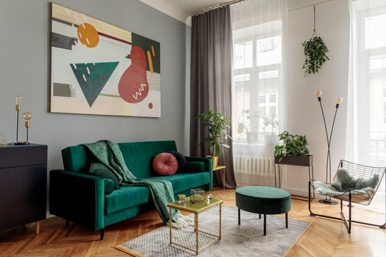 Luxury and stylish  interior with green velvet design sofa, armchair, tables and pouf. Tropical plants in design stand. Grey walls with abstract painting. Stylish decor of sitting room. Big windows.