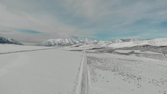 Aerial-Descending over snowy road running toward majestic snow-covered mountain in the distance under blue sky with high wispy clouds