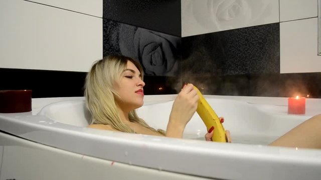 An attractive woman sits in the bathroom and brushes the big yellow banana and starts with the pleasure of eating it.  A girl eats a banana after a hard day