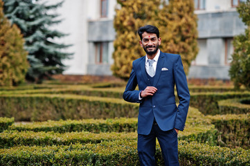 Stylish beard indian man with bindi on forehead, wear on blue suit posed outdoor against green bushes at  park.