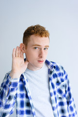 Curious young man holding hand near ear. Attractive guy looking at camera. News concept. Isolated front view on white background.